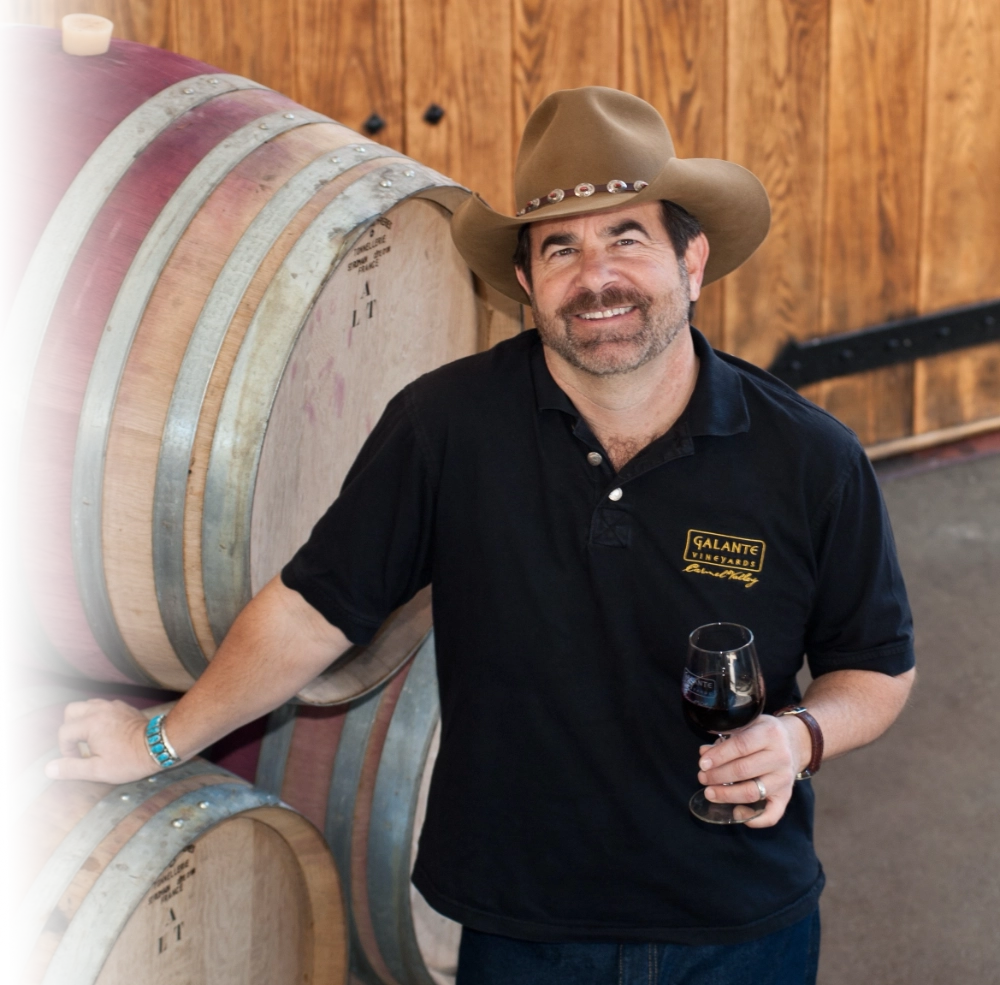 Man in a cowboy hat holding a glass of red wine, standing by wine barrels and smiling at the camera.