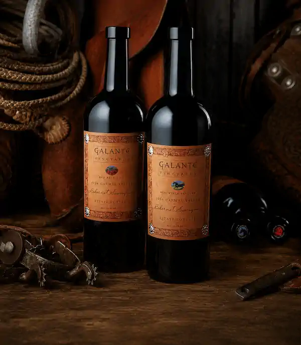 Two bottles of Galante Vineyards wine on a rustic table with a lasso, spurs, and saddle in the background.