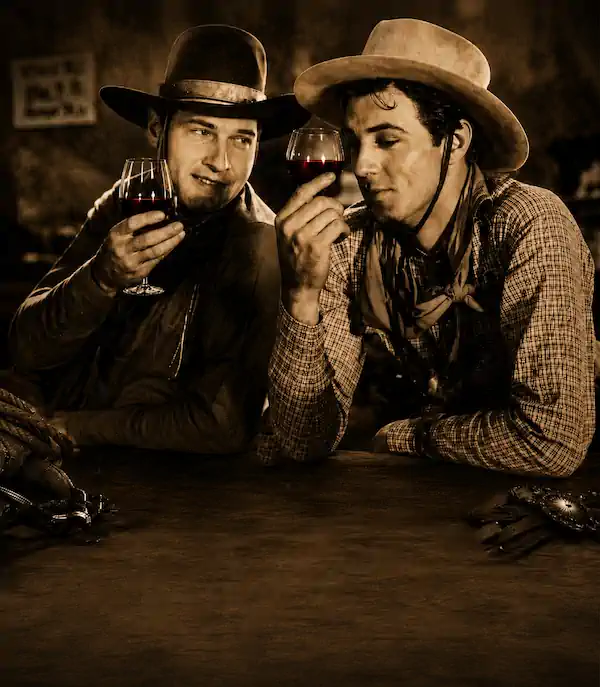 Two cowboys in hats sit at a bar, smiling and sipping red wine from glasses.