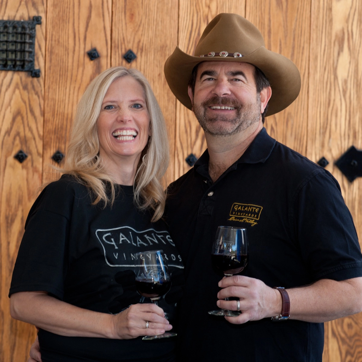 Smiling couple holding wine glasses, standing together in front of a wooden door, wearing Galante Vineyards shirts.