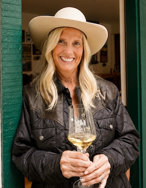 Smiling woman in a wide-brimmed hat holding a glass of white wine, standing by a green door.