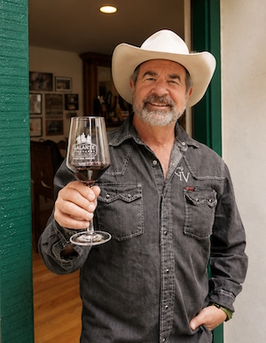 Man in cowboy hat smiles, holding a glass of red wine while standing in a doorway.