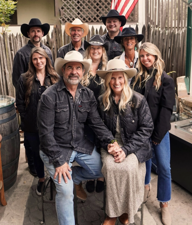 Nine people in cowboy hats and casual clothes pose and smile outdoors in front of a wooden fence and flag.