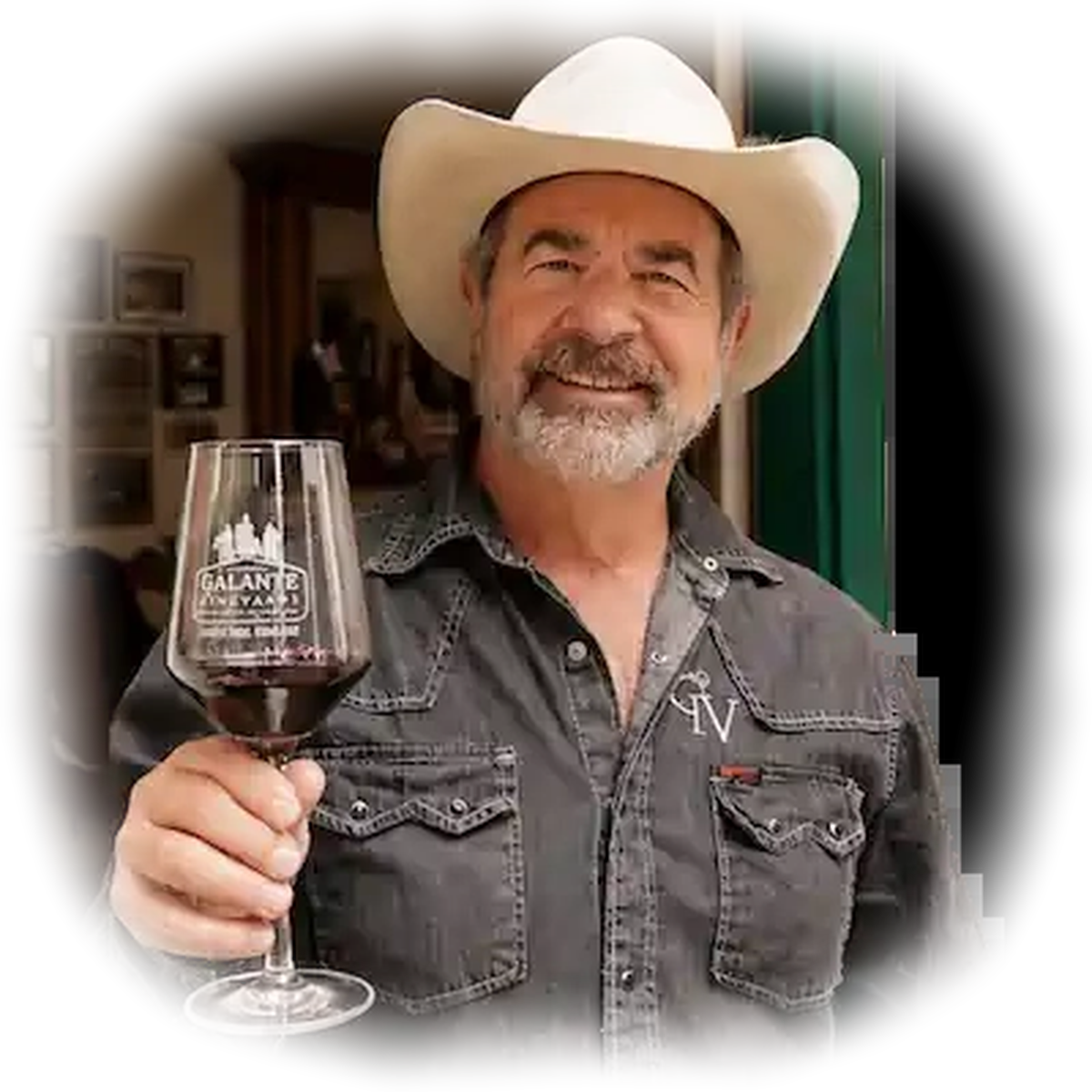 Smiling man in a cowboy hat holding a glass of red wine, wearing a dark shirt with white trim.