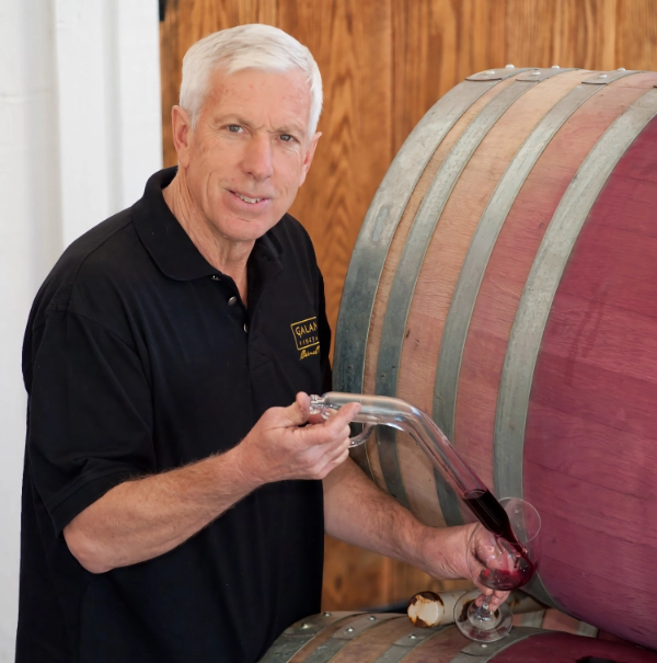 Man in black shirt pouring wine from a barrel into a glass using a wine thief, standing indoors.