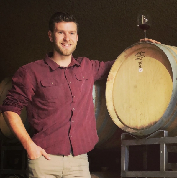 Man in maroon shirt stands by a wine barrel with a glass of red wine on top, smiling at the camera.