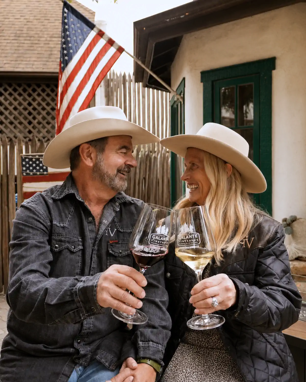 Two people in cowboy hats toast wine glasses and smile, sitting outside with an American flag in the background.